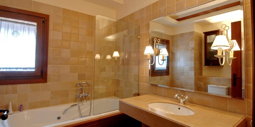 Un baño limpio y estilizado que presenta una gran bañera blanca con accesorios de cromo, un lavabo de encimera moderno con un grifo elegante y un espejo colgado en la pared. Azulejos beige cálidos adornan las paredes, y la luz natural entra a raudales a través de una pequeña ventana decorada con cortinas ligeras. Dos apliques de pared proporcionan una iluminación suave, mejorando la atmósfera acogedora, con un gabinete de madera oscura visible en el fondo.