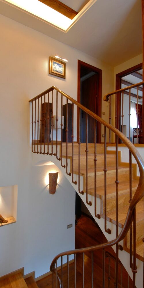 Escalera de madera curva con peldaños pulidos y barandillas de hierro que conduce a un nivel superior en un hogar luminoso. Una imagen enmarcada y un pequeño alcoba iluminada que exhibe una escultura decoran el espacio.