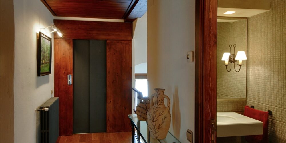 Un pasillo sofisticado que muestra un techo y suelo de madera, con una puerta de ascensor de madera oscura. A la izquierda, una mesa de cristal adornada con un jarrón tradicional que presenta diseños florales. El espacio está iluminado por una luz de pared, que resalta una pintura de paisaje enmarcada. En el fondo, se puede ver un baño moderno, equipado con accesorios contemporáneos, que incluyen un lavabo elegante y una toalla roja vibrante, realzado por la cálida iluminación de apliques de pared. Este diseño armoniza la elegancia clásica con la funcionalidad moderna.