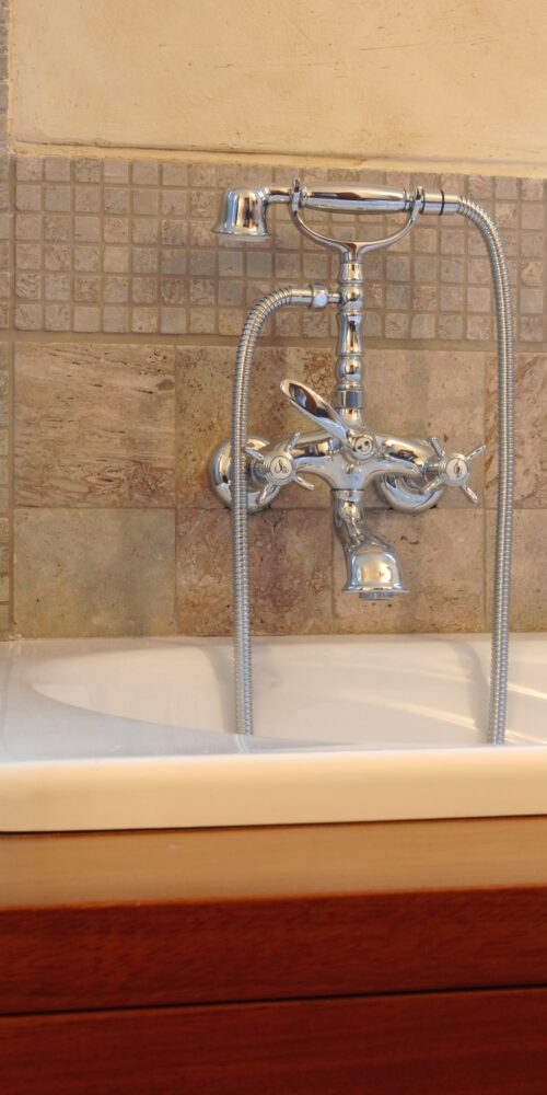 Vista en primer plano de un grifo de baño de estilo vintage con acabados en cromo, que cuenta con tres manijas para el control de temperatura y un rociador manual, montado en un fregadero rectangular blanco con un fondo de baldosas de piedra texturizada.
