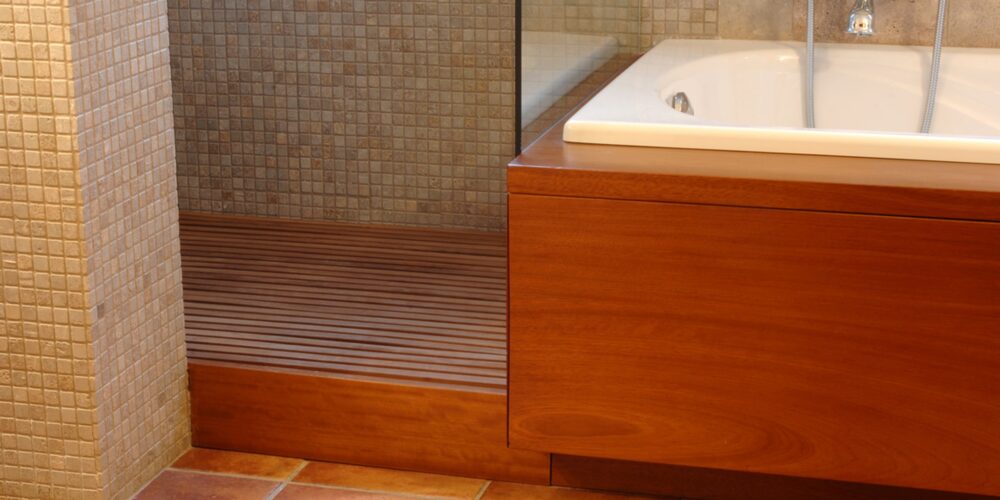 Interior de un baño contemporáneo que presenta un mueble de madera, una bañera blanca, un área de ducha tipo walk-in con paredes de azulejos y un suelo de madera listonada, iluminado por una suave luz natural.