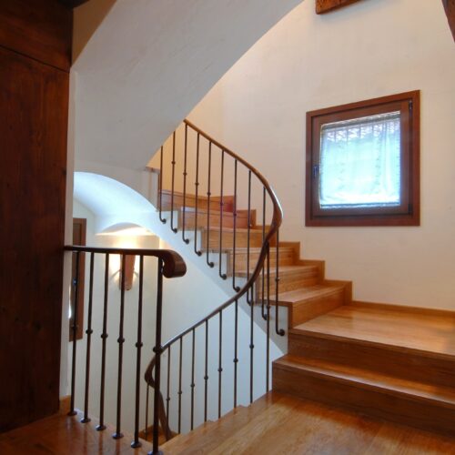 Vista interior de una elegante escalera de caracol con escalones de madera y un barandal de metal decorativo, iluminada por la luz natural que entra por una ventana cercana. Las paredes de color claro y el cálido suelo de madera realzan la atmósfera acogedora, enmarcada por vigas de madera rústica arriba.