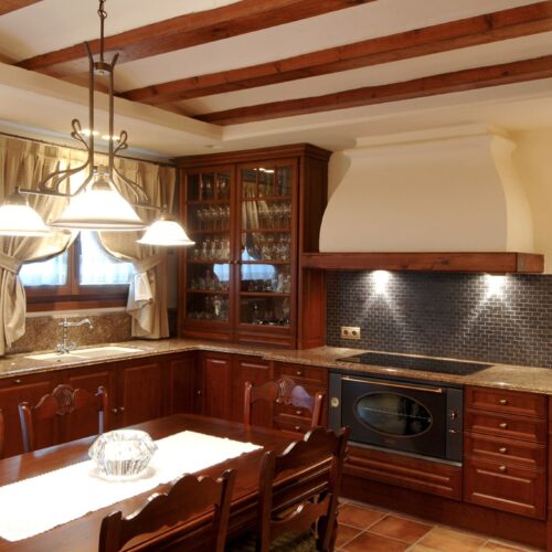 Una cocina acogedora que presenta muebles de madera, una espaciosa encimera de granito y una gran mesa de comedor con ocho sillas, iluminada por luces colgantes. Una gran ventana con cortinas deja entrar luz natural, realzando la atmósfera cálida. La cocina está equipada con un horno moderno y presenta un elegante backsplash decorativo hecho de azulejos oscuros. En los muebles se pueden ver cristalerías elegantemente exhibidas.