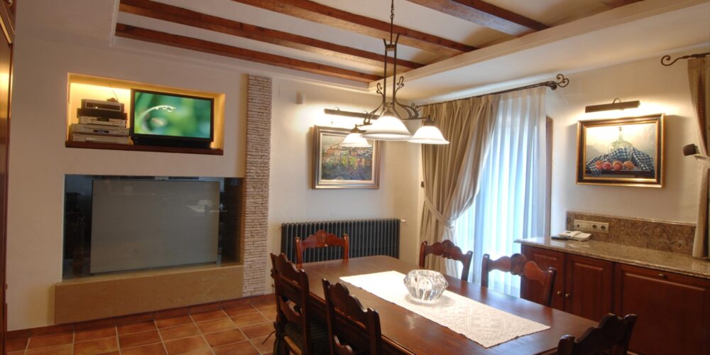 Una sala de comedor cálida y acogedora con una mesa de comedor de madera y sillas a juego, abundante luz natural, vigas de madera expuestas, un televisor de pantalla plana, una pared de piedra decorativa y acentos decorativos.