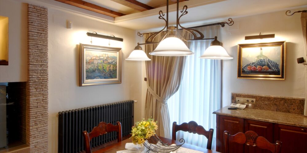 Un área de comedor cálida y acogedora con una mesa de madera elegantemente dispuesta con un centro de mesa floral, suave iluminación colgante, mantel de encaje, obras de arte enmarcadas y un radiador tradicional, creando un ambiente perfecto para reuniones.