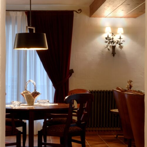 Una acogedora área de comedor con una mesa redonda de madera, decorada con vajilla elegante, una cesta decorativa y iluminada por una moderna lámpara colgante negra. Cortinas de un rico color burdeos y apliques de pared cálidos crean una atmósfera acogedora, complementada por una viga de madera rústica y una alfombra de área con patrón. Un radiador vintage y sillones mullidos son visibles en el fondo.