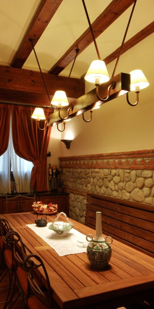 Un comedor cálido y acogedor con una mesa de madera rodeada de sillas elegantes, un lustre rústico con lámparas amarillas, vigas de madera ricas y cortinas suaves. Jarrones de cerámica decorativos adornan la mesa, con una pared de piedra al fondo y luz natural brillante filtrándose a través de la ventana.