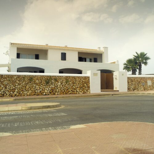 Casa moderna de dos pisos con paredes exteriores blancas, techo de tejas, persianas negras y entradas en arco, situada en un entorno suburbano sereno. Un muro de piedra rodea la propiedad, complementando la fachada. Un camino pavimentado y un paso peatonal conducen a una gran puerta de madera, mientras que una palmera se encuentra cerca, indicando un clima cálido.