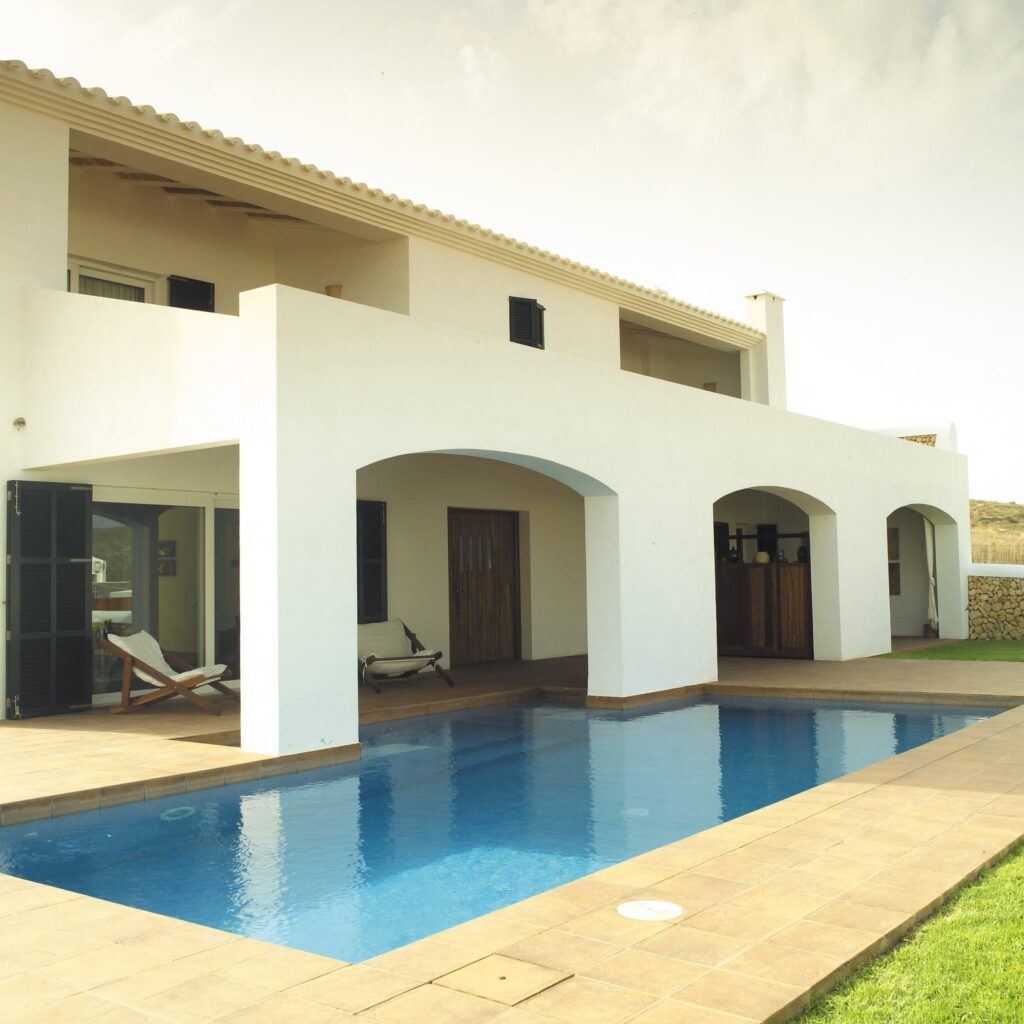 Chalet en Menorca: dos pisos con un elegante exterior blanco, rodeado de un exuberante césped verde. La villa cuenta con un amplio espacio exterior con una piscina, un patio de baldosas y tumbonas para relajarse, mientras que grandes arcos y puertas de vidrio iluminan el interior con luz natural.