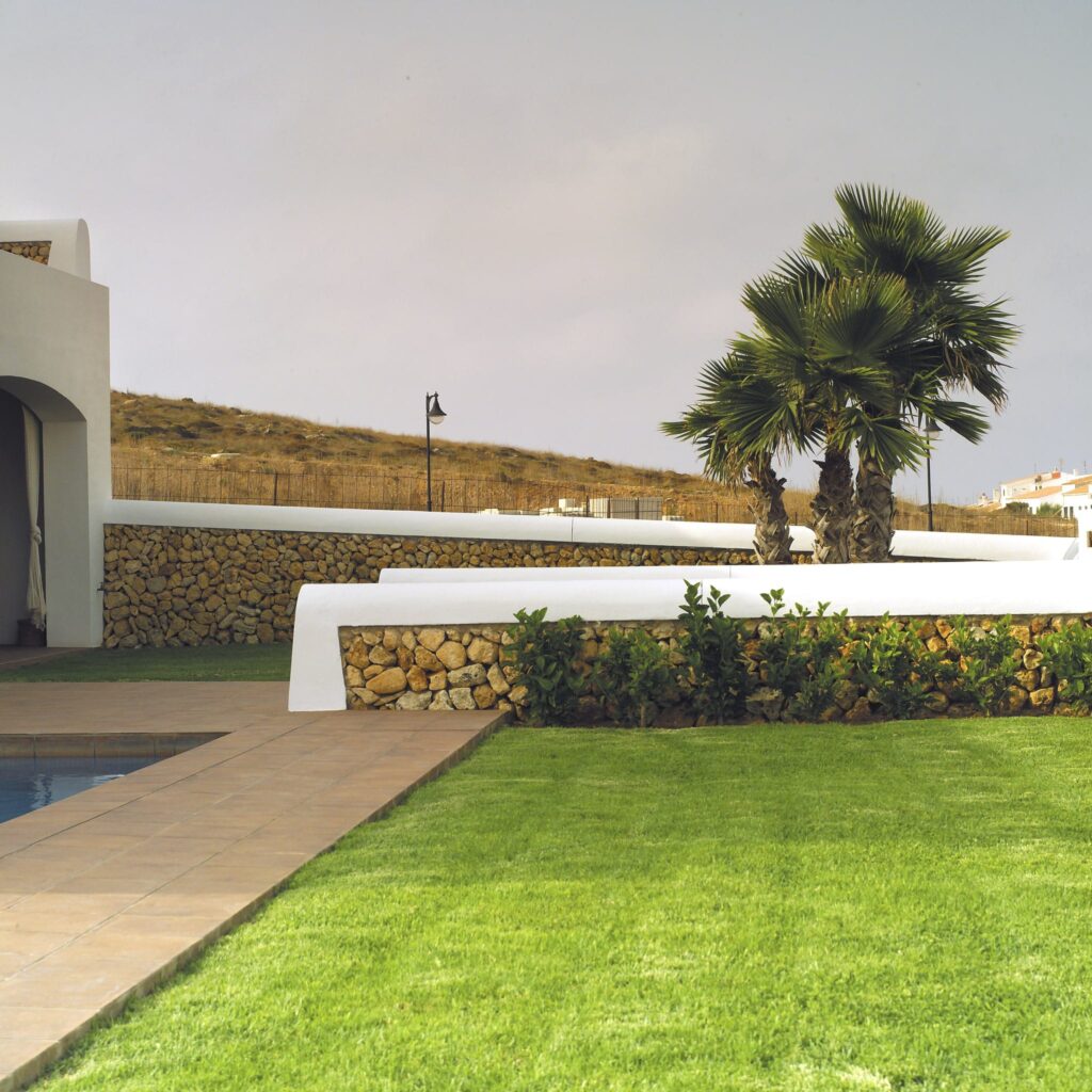 Paisaje al aire libre sereno con piscina y vibras tropicales Terrazas y jardines: Una hermosa área al aire libre con un césped verde exuberante, un sendero de baldosas que conduce a una piscina tranquila, rodeada de plantas bien cuidadas, un muro de piedra blanca, una característica rocosa natural, altas palmeras y una suave pendiente bajo un cielo nublado.