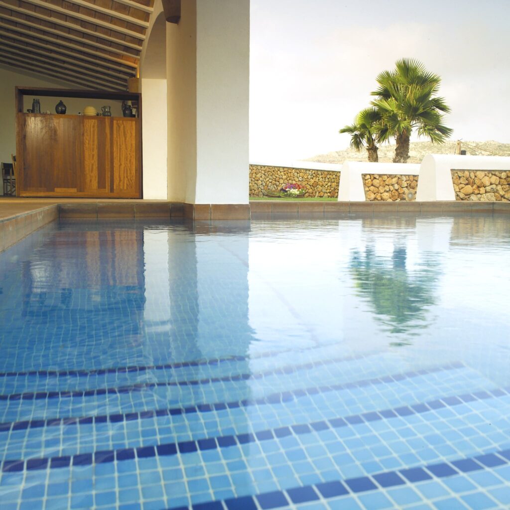 Hermosa piscina exterior rodeada de arquitectura moderna Una serena piscina exterior con agua azul clara que refleja las palmeras circundantes y el diseño arquitectónico moderno, con un bar de madera y paredes de piedra natural para un retiro vacacional tranquilo.