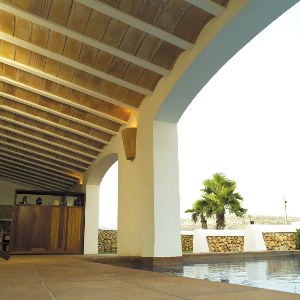 Elegante Interior Arquitectónico con Vigas de Madera y Serenas Vistas al Exterior Un acogedor interior arquitectónico con un techo de madera con vigas expuestas, un bar de madera con objetos decorativos, grandes arcos que conducen a un área exterior con palmeras, mampostería de piedra y agua de una piscina reflectante, creando una atmósfera serena.