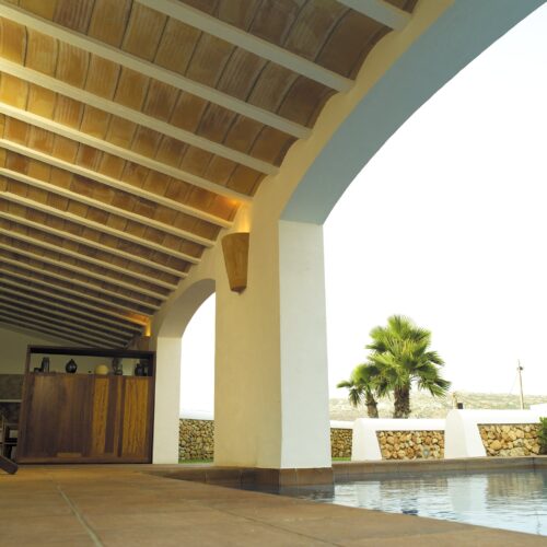 Un acogedor interior arquitectónico con un techo de madera con vigas expuestas, un bar de madera con objetos decorativos, grandes arcos que conducen a un área exterior con palmeras, mampostería de piedra y agua de una piscina reflectante, creando una atmósfera serena.
