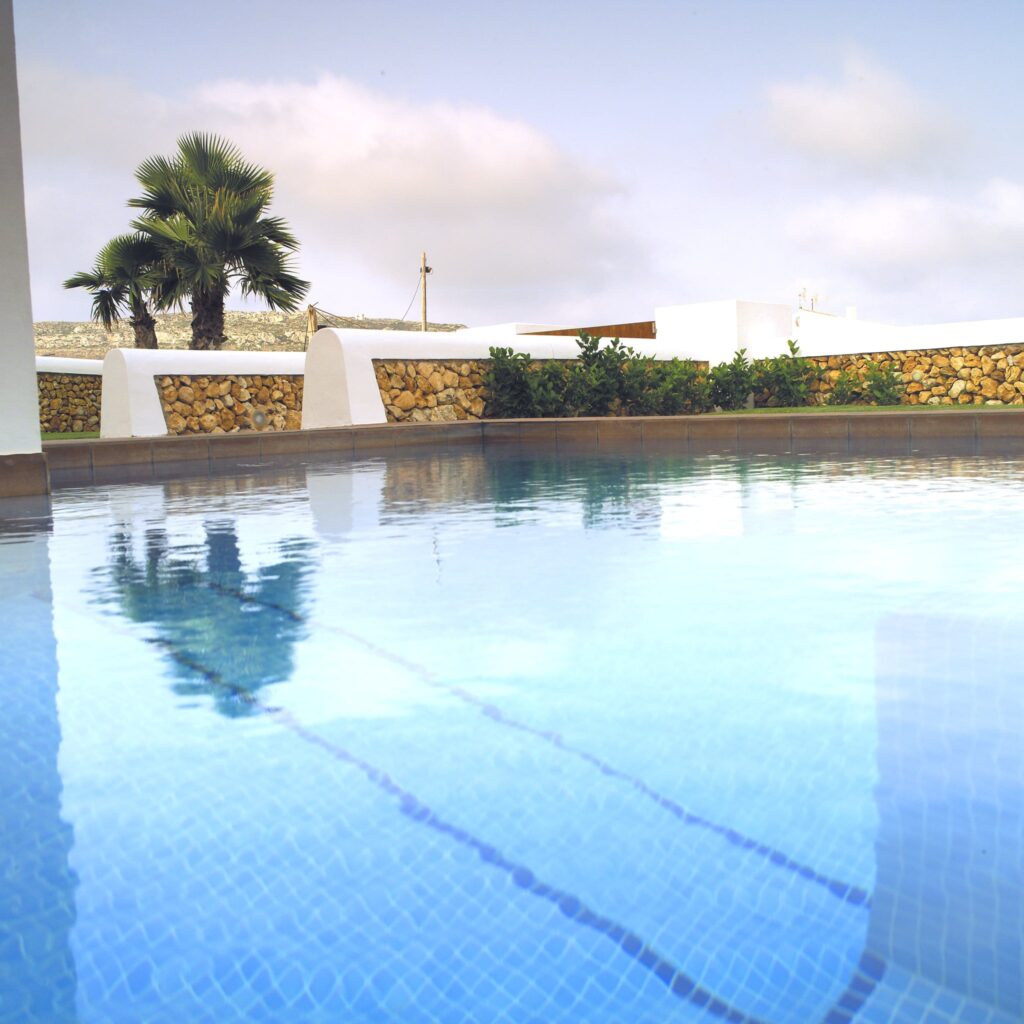 Oasis de Piscina Tranquila con Lujosas Palmeras Una escena serena de piscina mostrando agua azul cristalina reflejando un cielo nublado, rodeada de vibrantes palmeras verdes y elegante arquitectura blanca, enmarcada por una sofisticada pared de piedra.