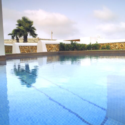 Una escena serena de piscina mostrando agua azul cristalina reflejando un cielo nublado, rodeada de vibrantes palmeras verdes y elegante arquitectura blanca, enmarcada por una sofisticada pared de piedra.