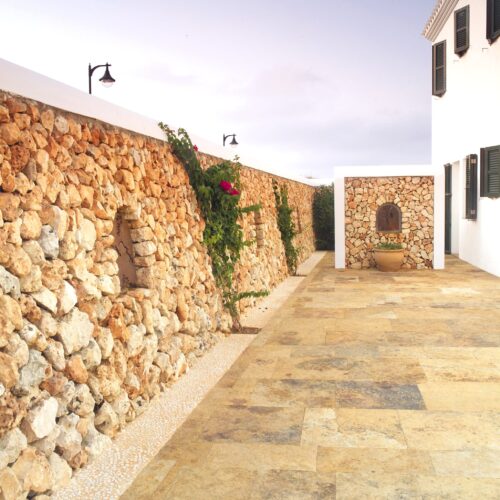 Un camino al aire libre tranquilo bordeado por muros de piedra de color claro adornados con plantas trepadoras y flores rosadas vibrantes, que conduce a una pequeña estructura blanca con características decorativas de piedra y una gran planta en maceta.