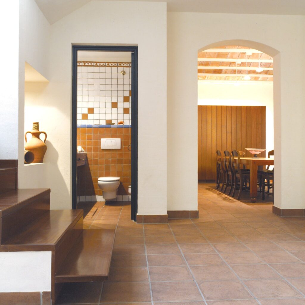 Muestra de Diseño Interior Moderno: Área Abierta con Vistas al Comedor y Baño Un espacio interior moderno que muestra una transición fluida entre un área abierta, un comedor con una larga mesa y sillas de madera oscura, y un baño con paredes de azulejos en tonos cálidos. La imagen presenta escaleras de madera que conducen hacia arriba, una maceta de cerámica en un alfeizar, y un arco que enmarca el área del comedor para un ambiente de vida armonioso.