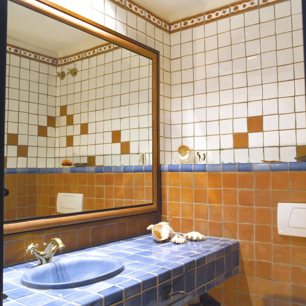 Baño Acogedor Inspirado en la Playa con Azulejos Vibrantes Un baño diseñado de manera única que presenta azulejos cuadrados en blanco, naranja y azul en las paredes, un gran espejo con marco de madera, un mostrador de azulejos azules y un lavamanos de porcelana con un grifo dorado, adornado con conchas decorativas.