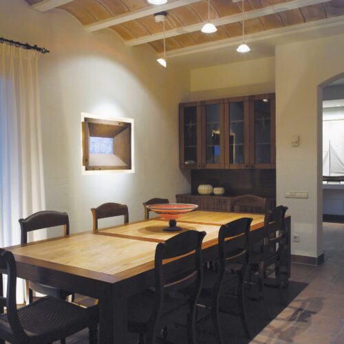 Un comedor cálido y acogedor que presenta una gran mesa de madera rodeada de sillas de madera negras, adornada con un cuenco decorativo. La suave iluminación ambiental resalta una obra de arte en la pared y un vitrina con puertas de vidrio. Las cortinas color crema caen sobre la ventana, permitiendo que la luz natural ilumine el suelo de baldosas.