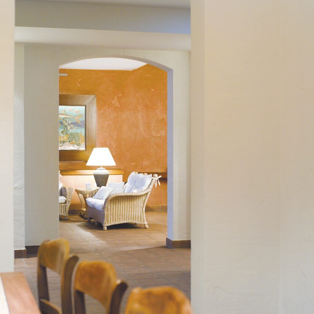 Área de salón acogedora con muebles de mimbre y acentos cálidos Vista interior de una acogedora área de salón que presenta muebles de mimbre de color claro con cojines blancos, iluminada por una lámpara de pie contra una pared de tonos naranjas cálidos, vista a través de un arco.
