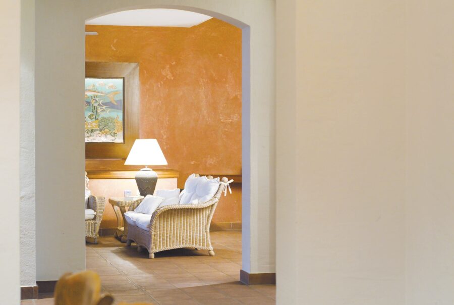 Vista interior de una acogedora área de salón que presenta muebles de mimbre de color claro con cojines blancos, iluminada por una lámpara de pie contra una pared de tonos naranjas cálidos, vista a través de un arco.