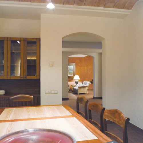 Vista interior de un comedor moderno que presenta una mesa de madera clara y sillas de madera oscura, que da a una sala de estar iluminada cálidamente con asientos acogedores y una lámpara, mostrando una mezcla de diseño contemporáneo y rústico.