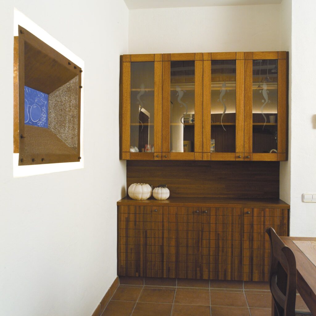 Interior Minimalista Moderno con Gabinete de Madera y Arte Abstracto Brillante interior moderno que muestra un gabinete de madera con puertas de vidrio y base de madera maciza, acompañado de dos calabazas blancas decorativas. Una pieza de arte abstracto única, texturada en tonos marrones y azules, cuelga en la pared, sobre un suelo de baldosas, con una mesa de comedor de madera en la esquina.