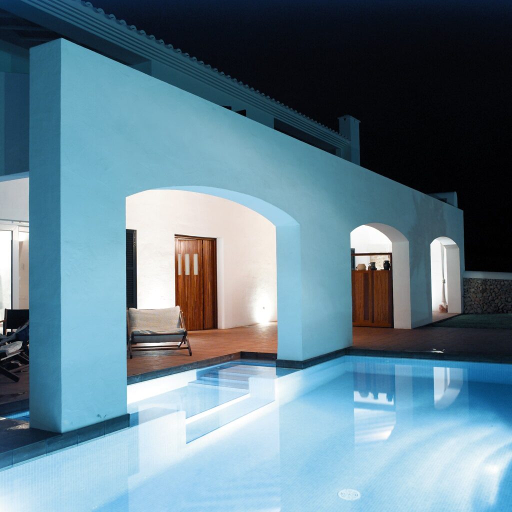 Arquitectura Moderna Serena de Noche con Piscina Reflectante Vista nocturna de un edificio de arquitectura moderna con puertas arqueadas y suave iluminación, con una tranquila piscina en primer plano que refleja luz azul.