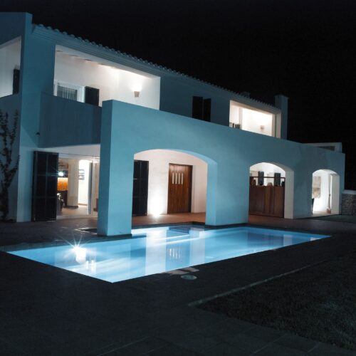 Una impresionante villa moderna de dos pisos con un exterior blanco, puertas de madera oscura y elegantes arcos, rodeada de un acogedor área de piscina por la noche. La piscina está suavemente iluminada, realzando la serena atmósfera del lujoso espacio al aire libre.
