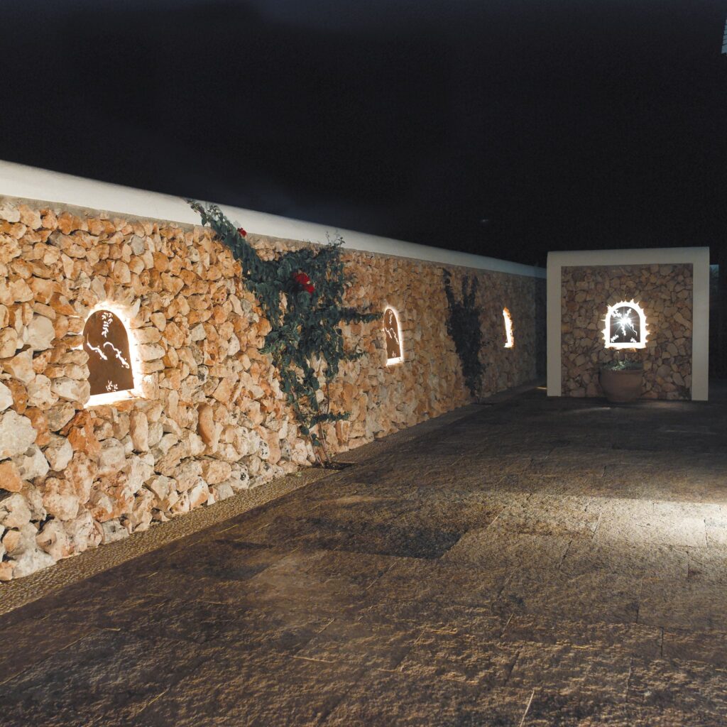 Pared de Piedra Rústica Tranquila con Iluminación Suave y Vides por la Noche Una pared de piedra rústica iluminada por luces suaves en recesos arqueados, con vides verdes trepadoras y flores rojas, situada sobre un pavimento de piedra oscura por la noche.