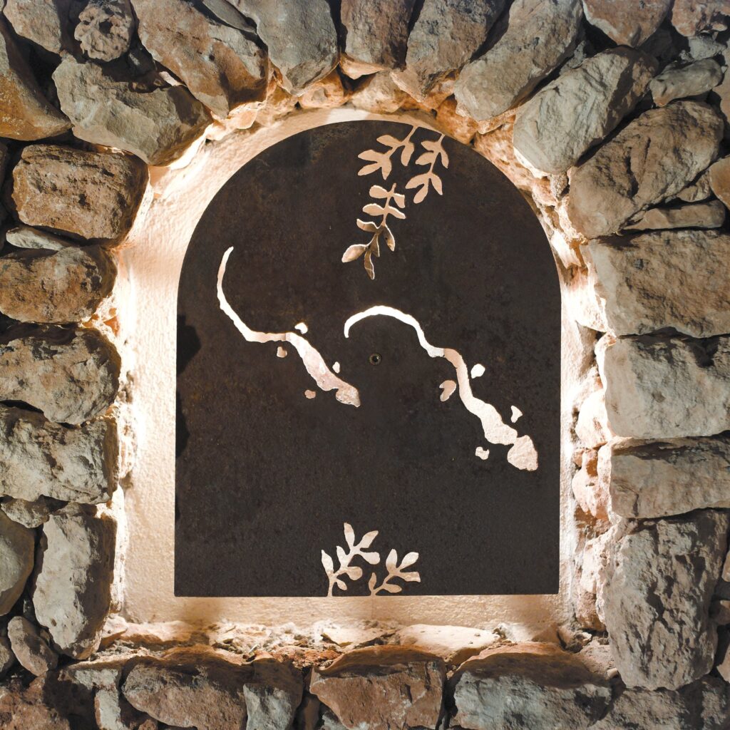Arco Decorativo Iluminado con Diseño de Panel Intrincado Un arco decorativo de piedra natural que presenta un panel oscuro con diseños recortados detallados que asemejan líneas fluidas y hojas, iluminado desde atrás. El arco está rodeado de piedras de varios tonos de gris y beige, creando una atmósfera rústica y cálida.