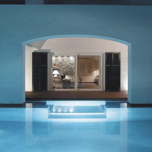 Una vista serena de un área interior moderna enmarcada por un elegante arco, con grandes puertas de vidrio que dan a una sala de estar estilosa llena de muebles acogedores y papel tapiz con patrones. La tranquila piscina en primer plano refleja suaves tonos azules, creando una atmósfera pacífica y mostrando la fusión de la comodidad interior con el encanto exterior.