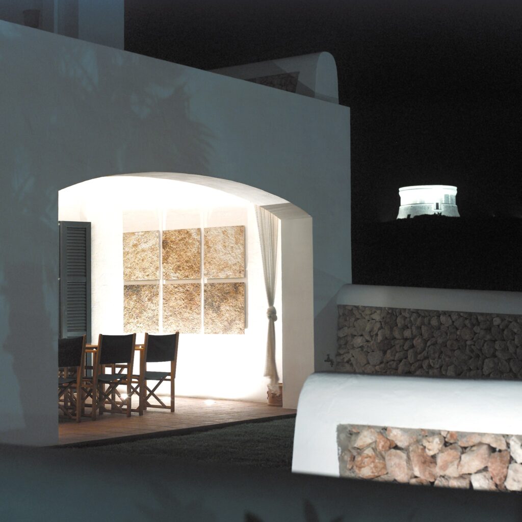 Elegante Área de Comedor al Aire Libre con Entrada Arqueada Arquitectónica Un área de comedor al aire libre de diseño hermoso que presenta una estructura blanca con una entrada arqueada, grandes paneles de pared texturizados, sillas de madera con cojines negros y una suave cortina drapeada, enmarcada por un tranquilo cielo nocturno con una estructura luminosa en la colina de fondo.