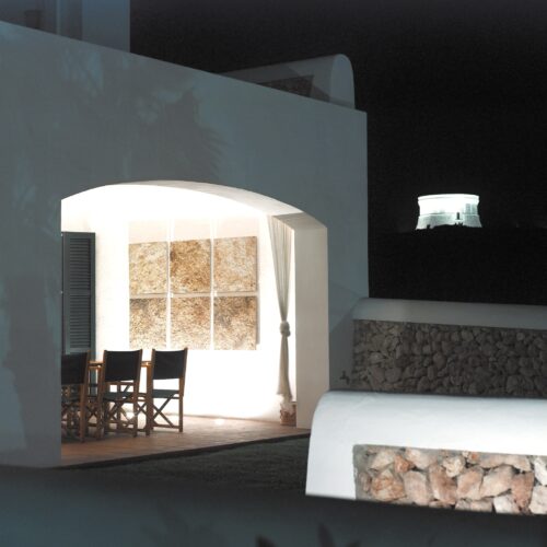 Un área de comedor al aire libre de diseño hermoso que presenta una estructura blanca con una entrada arqueada, grandes paneles de pared texturizados, sillas de madera con cojines negros y una suave cortina drapeada, enmarcada por un tranquilo cielo nocturno con una estructura luminosa en la colina de fondo.