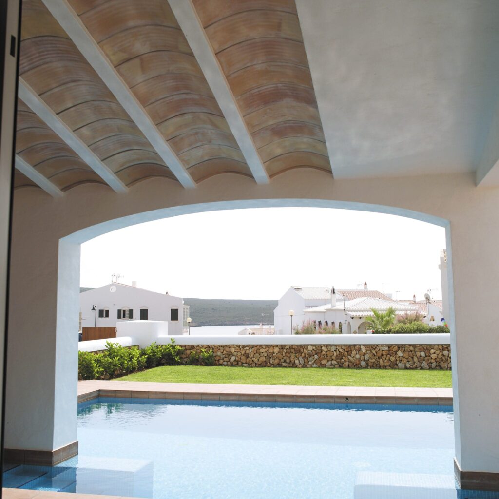 Escena exterior tranquila con piscina vista a través de una ventana arqueada Una serena vista exterior a través de una ventana arqueada que presenta una piscina con agua azul clara, bordeada por un patio de piedra y rodeada de hierba verde exuberante y arbustos recortados. En el fondo, edificios blancos con techos de terracota complementan la atmósfera pacífica, todo bajo una estructura con elegantes vigas de madera.
