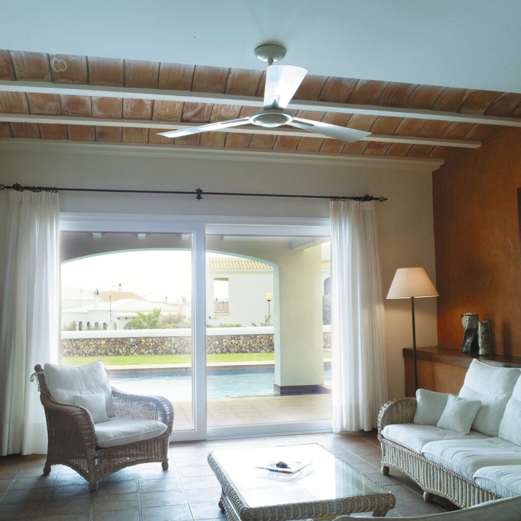 Sala de Estar Brillante y Aireada con Acceso a la Piscina Exterior Una amplia y luminosa área de estar que muestra una puerta corredera de cristal que conduce a una zona de piscina exterior. La sala cuenta con un sofá y sillas de mimbre de color claro con cojines blancos suaves, un ventilador de techo moderno y suaves cortinas blancas que crean una atmósfera serena.