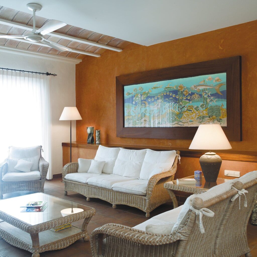 Sala Acogedora con Sofá de Ratan y Arte Submarino Una encantadora sala de estar que exhibe un juego de sofá de ratán adornado con cojines blancos, complementado por una mesa de café de vidrio. El espacio cuenta con una pintura decorativa en la pared con vibrantes escenas submarinas, realzando la atmósfera. Una lámpara alta y jarras elegantemente dispuestas están cerca de una gran ventana vestida con cortinas ligeras. Las cálidas paredes naranjas contribuyen a la atmósfera acogedora, creando un entorno bien iluminado y decorado con buen gusto, ideal para la relajación.