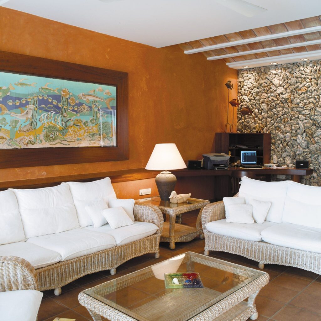 Sala de Estar Acogedora con Asientos de Mimbre y Acentos Naranjas Vibrantes Una sala de estar cálida y acogedora que presenta dos sillas de mimbre blancas tapizadas adornadas con coloridos cojines, una mesa de café de mimbre a juego, una obra de arte decorativa con temática de peces sobre el sofá, y una lámpara elegante que proporciona luz suave al lado de una pared de piedra natural. También se puede ver un escritorio de madera con una computadora, creando un espacio funcional adecuado para la relajación y reuniones.