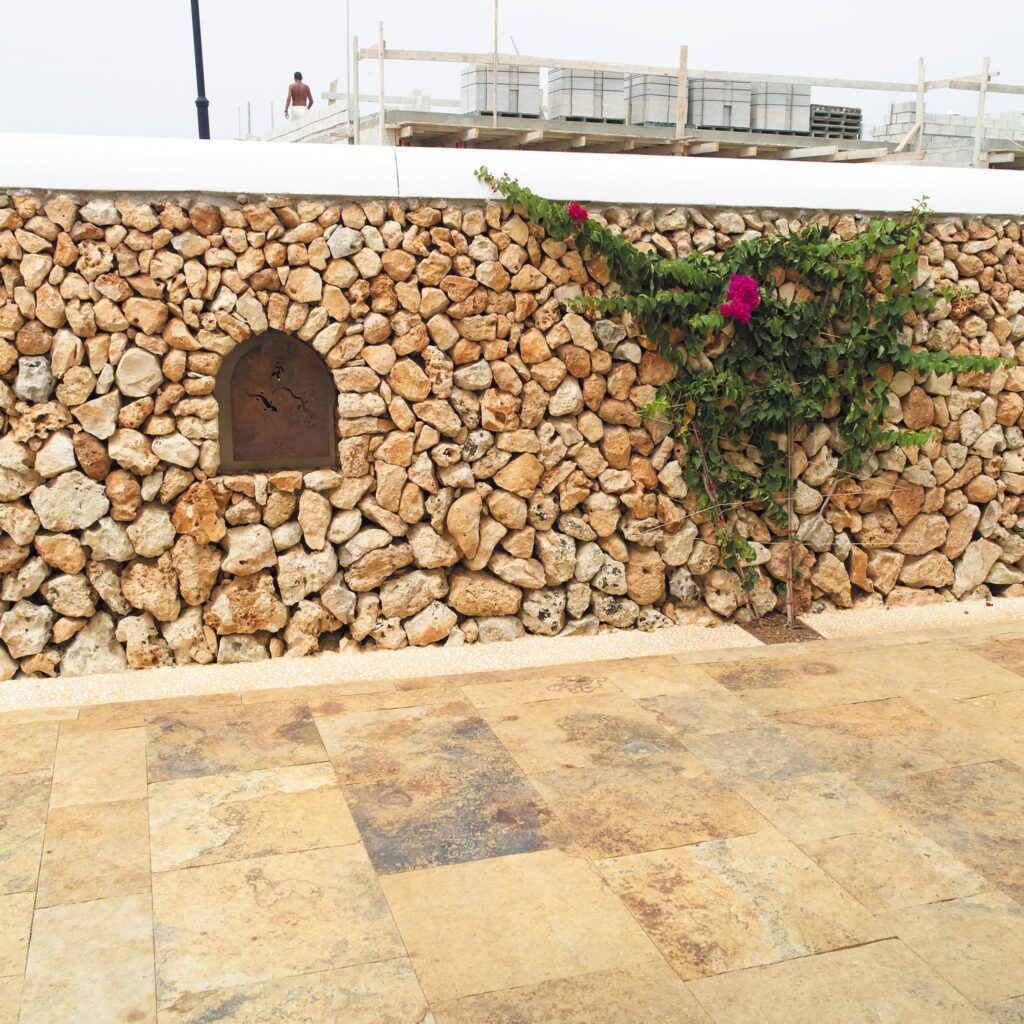Encantadora Pared de Piedra Rústica con Vides Coloridas Una pared de piedra rústica hecha de varias piedras en tonos beige y marrón claro, que presenta una pequeña nicho arqueado y una vid verde frondosa con vibrantes flores rosas creciendo a su lado. El suelo está pavimentado con grandes losas de piedra rectangulares en tonos terrosos, y en el fondo aparecen materiales de construcción, indicando una renovación.