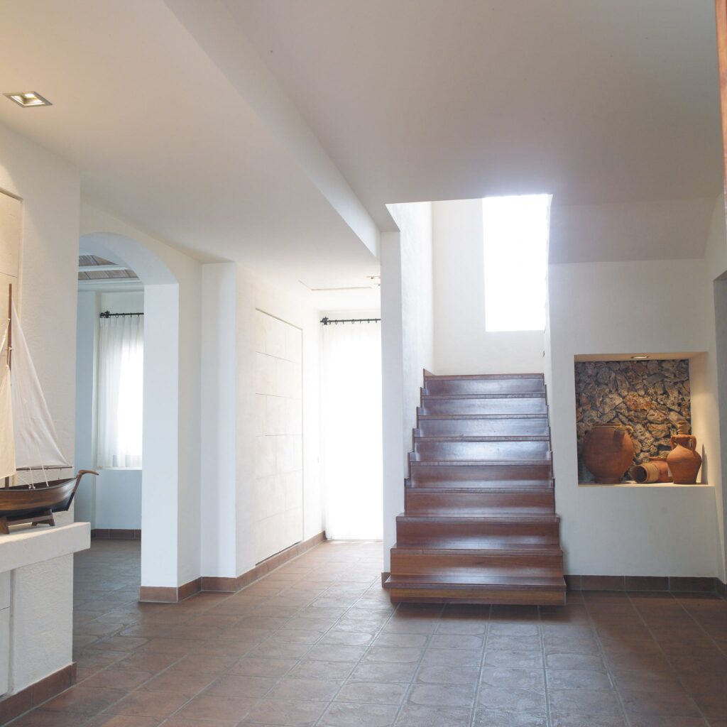 Casa Moderna Brillante y Espaciosa con Escalera de Madera Un interior de hogar moderno que presenta una escalera de madera, grandes ventanas que permiten la entrada de luz natural, paredes blancas, un modelo decorativo de velero en un estante, macetas de arcilla contra un fondo de piedra y suelos de baldosas cuadradas en tonos cálidos.