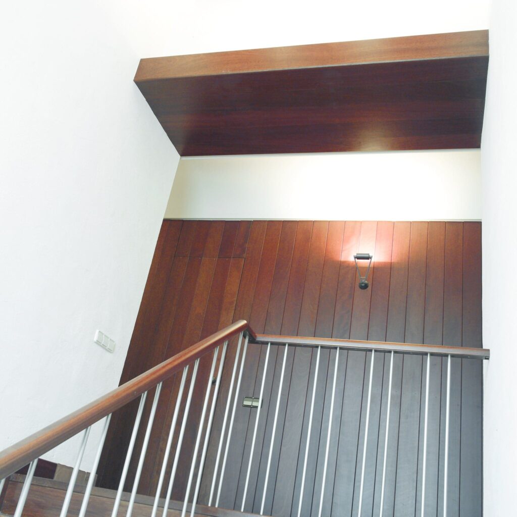 Escalera interior moderna con pasamanos de madera y balaustres metálicos Una escalera interior moderna y bien iluminada que muestra un pasamanos de madera y balaustres metálicos verticales contra paredes pintadas de blanco. La escalera está destacada por una pared revestida de madera y una simple lámpara de pared. La saliente de madera oscura en el techo agrega un toque contemporáneo a la estética.