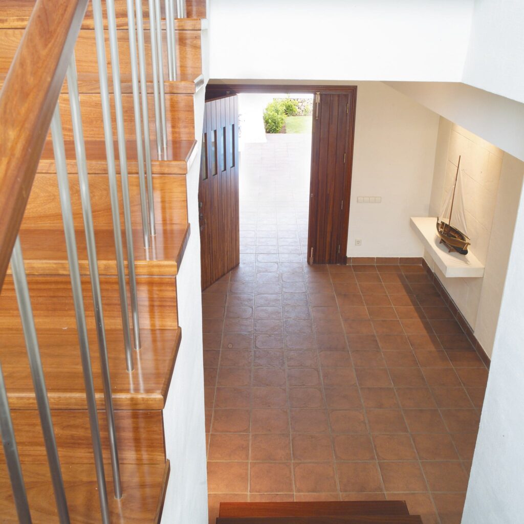 Vestíbulo de Escalera con Luz Natural y Exhibición de Barcos a Escala Vista superior de una escalera con escalones de madera y barandillas de metal que conducen a un amplio vestíbulo con un suelo de baldosas en tonos tierra y puertas dobles. Una estantería estrecha en la pared exhibe un pequeño modelo de barco, iluminado por la luz natural.