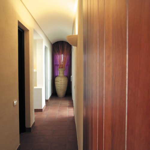Un pasillo interior sereno y elegante con paredes lisas de color claro, paneles de madera ricos, un jarrón decorativo alto con ramas secas y cortinas moradas, complementado por un suelo de baldosas en tonos terracota.