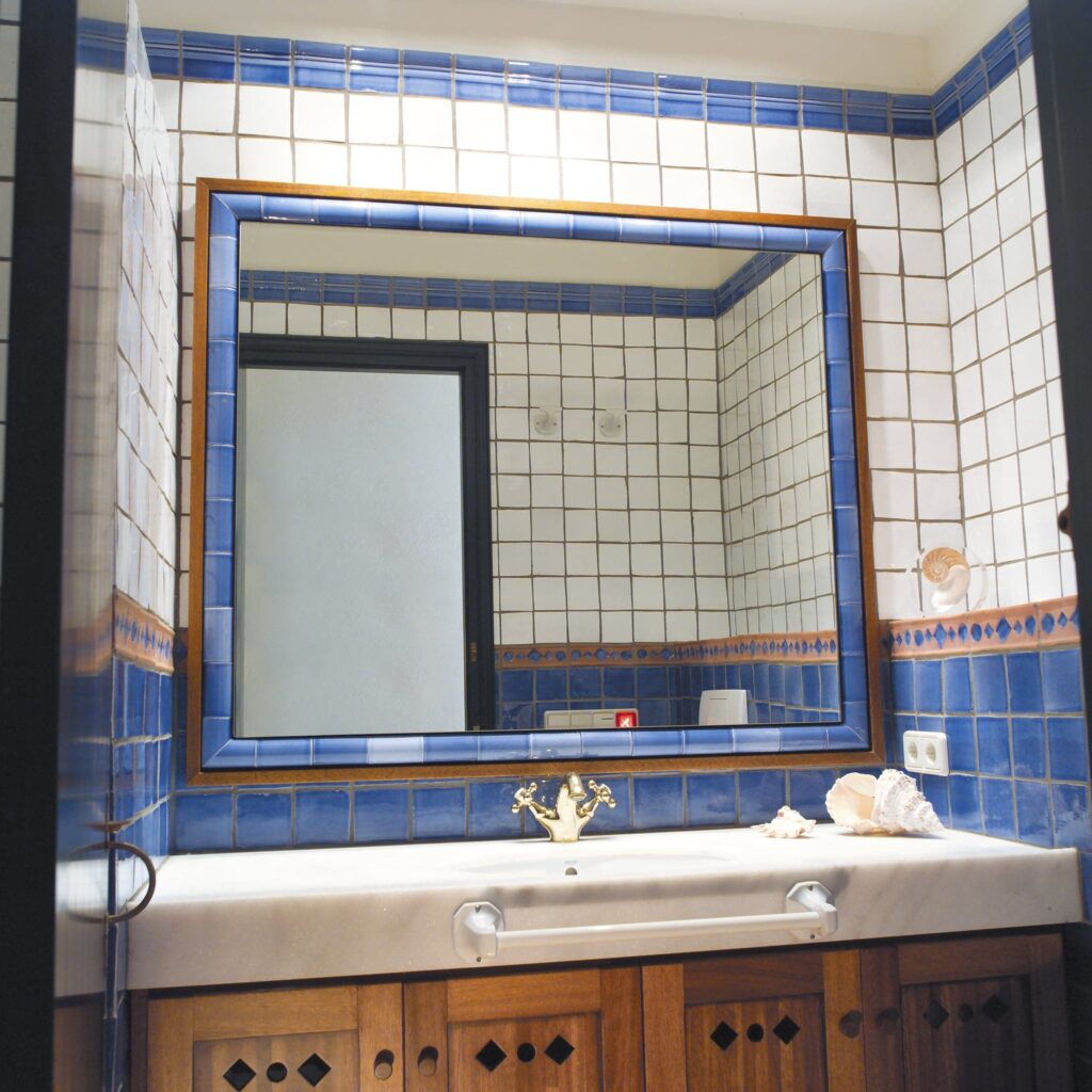 Baño Elegante con Azulejos Azules y Blancos y Decoración Vintage Un baño sereno con paredes de azulejos azules y blancos, un gran espejo rectangular con marco de madera, una encimera beige con un grifo dorado vintage y dos conchas decorativas. El armario de madera debajo de la encimera presenta intrincados recortes decorativos, y la luz natural inunda el espacio, enfatizando el diseño armonioso.
