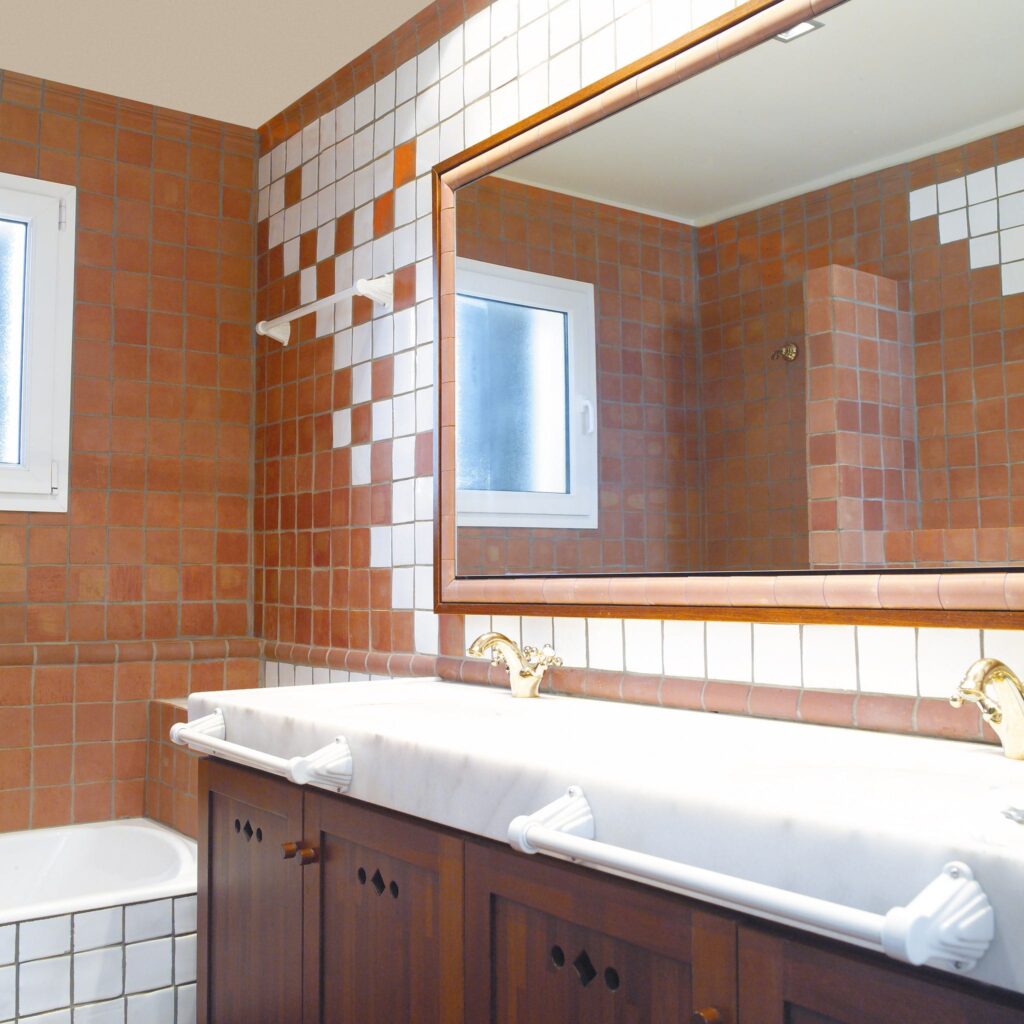 Baño Interior Estiloso con Azulejos de Terracota y Blancos Interior moderno de baño que muestra azulejos cuadrados de terracota y blancos en las paredes, un tocador de madera con una encimera de mármol, grifos de bronce, un gran espejo enmarcado y una ventana que permite que la luz natural llene el espacio.