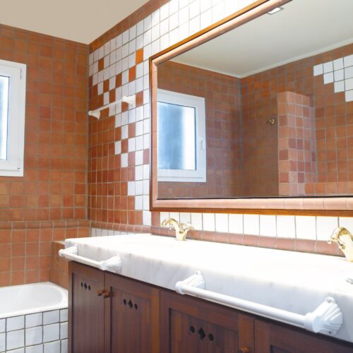Interior moderno de baño que muestra azulejos cuadrados de terracota y blancos en las paredes, un tocador de madera con una encimera de mármol, grifos de bronce, un gran espejo enmarcado y una ventana que permite que la luz natural llene el espacio.