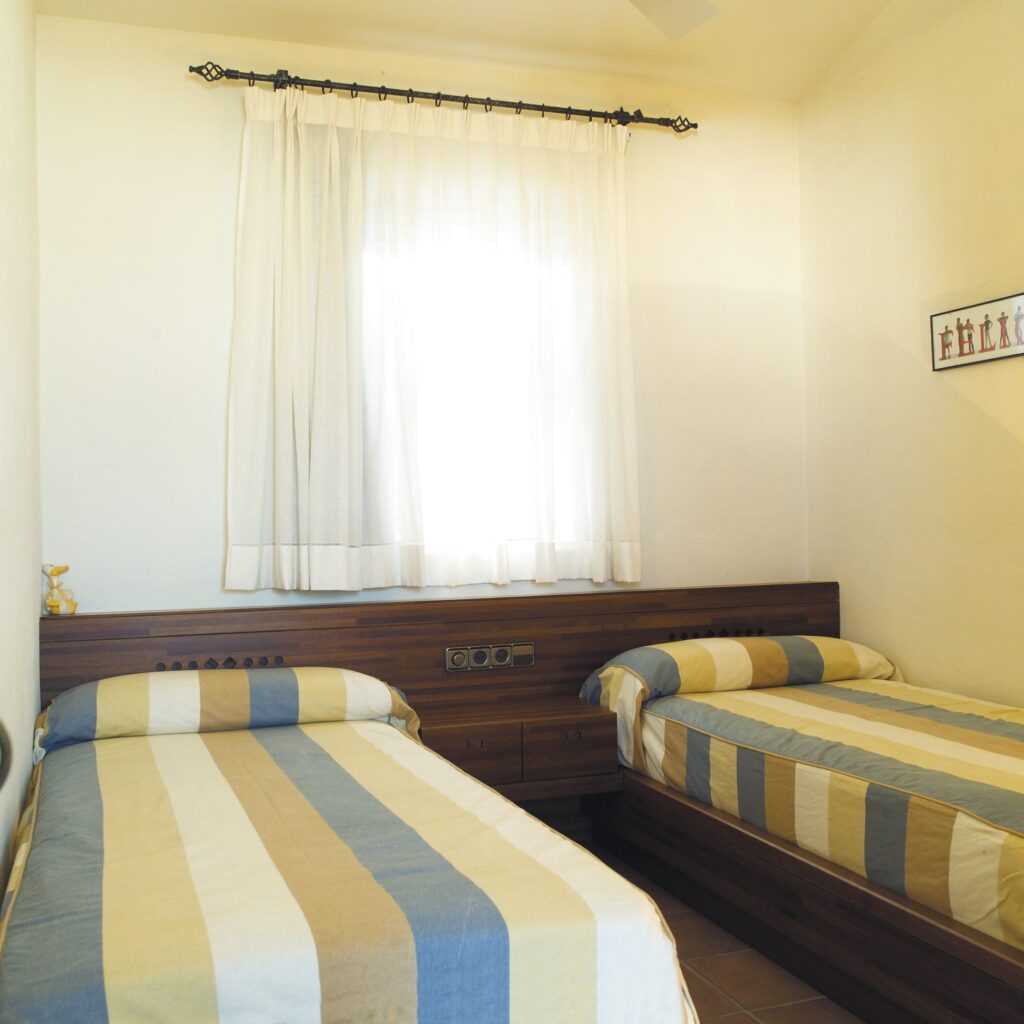 Acogedora Pequeña Habitación con Camas Individuales y Decoración Moderna Una pequeña habitación acogedora que presenta dos camas individuales con ropa de cama a rayas en azul y beige, ubicadas contra un cabecero de madera con almacenamiento integrado. La luz natural llena la habitación a través de una ventana con cortinas blancas translúcidas, realzando la atmósfera acogedora.