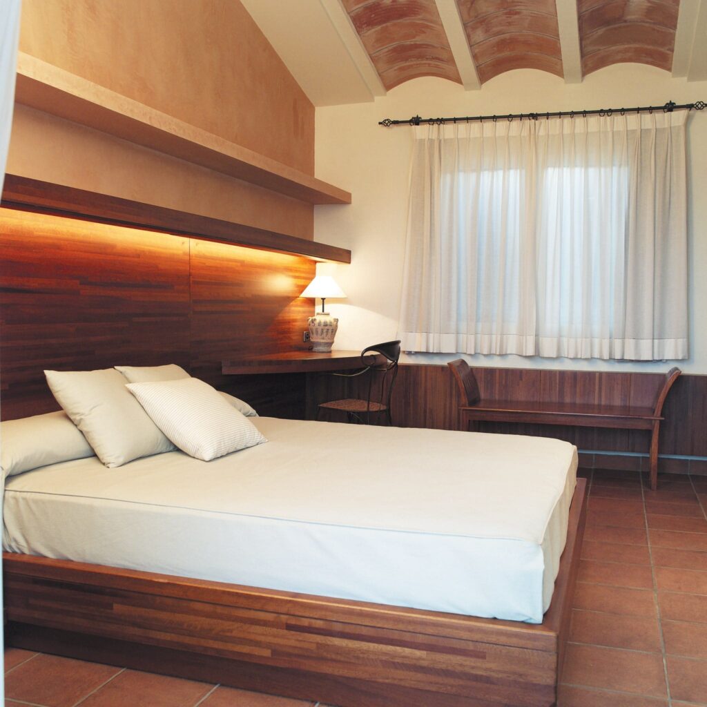 Dormitorio Moderno Minimalista con Estética Acogedora Un dormitorio minimalista con una cama bien hecha con ropa de cama de colores claros y una almohada estampada, un cabecero de madera cálido con iluminación integrada, luz natural filtrándose a través de cortinas ligeras, y un banco de madera sencillo contra la pared.