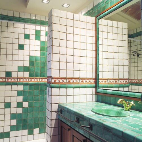 Baño brillante que presenta azulejos de rayas verde y blanco brillantes, una encimera de azulejos verdes a juego con lavabo integrado y un gran espejo enmarcado en azulejos verdes, iluminado por luz empotrada.