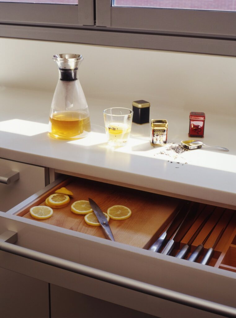 Escena de Cocina Serena con Bebida Refrescante de Limón y Especias Una cocina brillante y minimalista que presenta una jarra de vidrio con líquido amarillo, un vaso con una bebida similar y frascos de especias. Un cajón abierto muestra una tabla de cortar de madera con rodajas de limón y un cuchillo, junto a utensilios ordenadamente dispuestos.